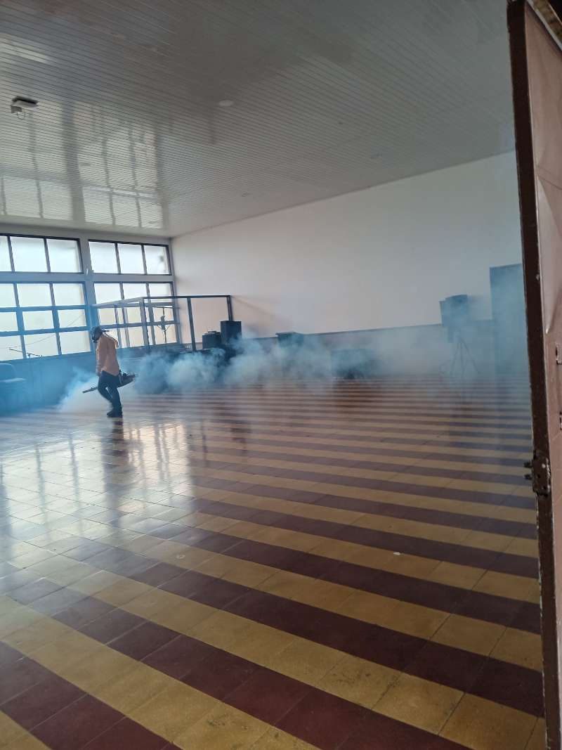 Técnico de EcoPlagas nebulizando un salón institucional amplio con niebla visible